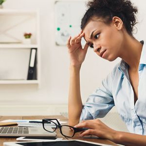 Deadline stress concept - sad african-american business woman sitting at desktop in office, working on laptop and holding hand on head. Hard working day, copy space